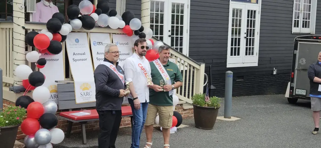 Three men with Captain sashes pose with trophies beneath a balloon arch at an outdoor awards ceremony next to sponsor boards.